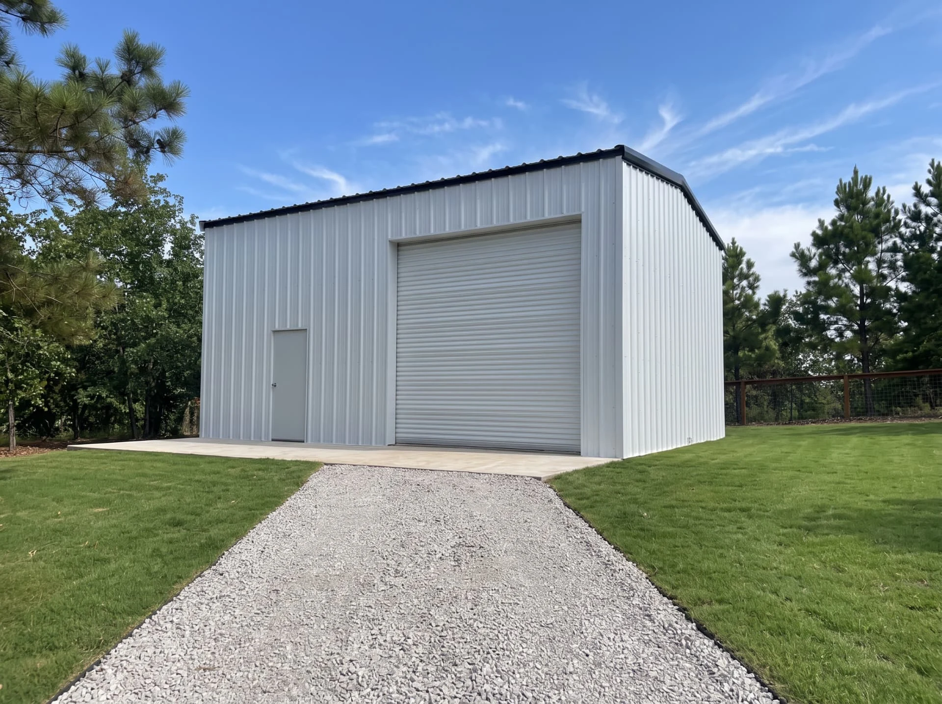 White metal shop building with roll-up door and gravel drive surrounded by pines